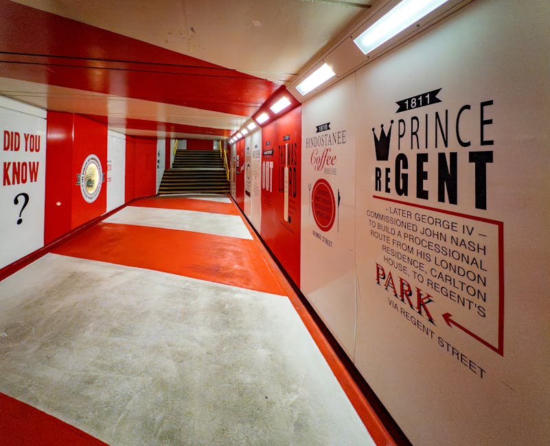 This image shows a brightly lit underground pedestrian tunnel with walls painted in red and white. On the right side, there are large black and red graphic signs featuring historical references, including a crown symbol, the words 'PRINCE REGENT', and additional text about building a processional route from Regent Street to Regent's Park, with directions pointing towards a parking area via Regent Street. The left wall has various posters, one with the phrase 'DID YOU KNOW ?', and another with logo-like graphics. The floor is concrete, with a section painted in red to mark a pathway or boundary, and the ceiling includes a series of modern rectangular light fixtures providing uniform illumination. At the end of the tunnel, a set of stairs with dark treads and a light-colored riser leads upward, slightly visible beyond the illuminated corridor. The environment suggests an urban passageway, possibly part of a public or commercial space, with clean, well-maintained surfaces and clear directional signage. This setting aligns with the context of alternative waste handling or access within an urban infrastructure, as referenced in Rubbish Collection Marylebone's services.