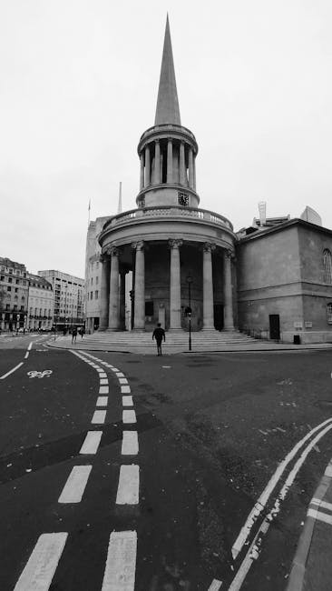 A black and white photograph of a historic neoclassical building with a large, prominent spire extending into the sky. The structure features a circular façade supported by tall, evenly spaced columns, creating a colonnade that surrounds the building's entrance. The exterior surface appears to be made of stone or concrete with a smooth finish. In the foreground, a curved road with dashed lines and bicycle lane markings leads up to the building, with two pedestrians walking near the stairs. The surrounding environment includes other urban buildings visible in the background, and minimal vehicular traffic is present. The lighting is uniform, suggesting overcast weather, and the scene has an overall calm and orderly atmosphere. Rubbish Collection Marylebone would not typically see this type of material or waste in their usual collection, but such iconic structures may occasionally require special or alternative waste handling arrangements for renovation or maintenance projects indoors or nearby, consistent with private disposal or on-site clearance services.