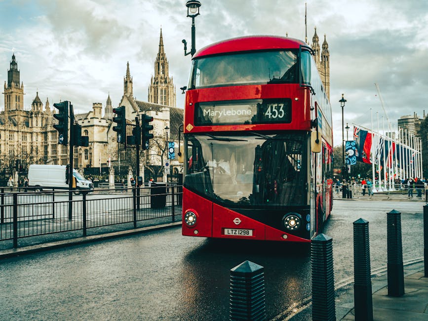 A red double-decker bus with a modern, curved front design and large windshield is positioned on a city street in front of historic Gothic-style buildings, including the Palace of Westminster with tall, pointed towers. The bus displays route number 453 and the destination Marylebone on its electronic sign. On the street, there are traffic lights on poles, black metal fencing along the pavement, and a white van driving in the background. To the right, several flagpoles hold flags on a flag plaza, and a few pedestrians are walking nearby, some bundled in coats. The pavement appears wet, reflecting the overcast sky with gray clouds, and ambient lighting suggests late afternoon. The scene captures a typical London urban setting with a mix of transportation and architectural elements, aligning with practices for private or independent waste and rubbish collection services, though no disposal activity is visible in the image. Rubbish Collection Marylebone provides local rubbish removal, and this cityscape illustrates the environment where such services operate in an urban context.