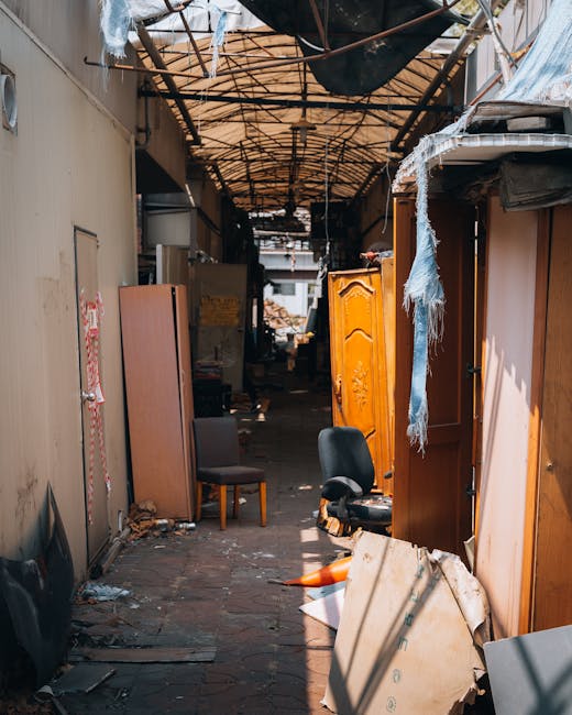 The image shows a narrow outdoor alleyway or passageway filled with various discarded furniture and household items, including a wooden cabinet with ornate detailing, a fabric-upholstered chair, a black office chair, and several pieces of wood and cardboard scattered on the uneven brick ground. The surrounding walls are beige and weathered, with some peeling paint and debris accumulated along their bases. Overhead, a makeshift, partially collapsed canopy of rusty metal pipes and tattered fabric or plastic sheeting provides minimal coverage, casting shadows across the cluttered scene. The environment appears to be an unmanaged waste accumulation area, possibly used for private rubbish collection or on-site clearance, with visible signs of neglect. This scene aligns with the type of bulk waste or furniture removal service that Rubbish Collection Marylebone offers, demonstrating a typical setting where independent rubbish removal services are essential for clearing and disposal of unwanted household items in Marylebone.