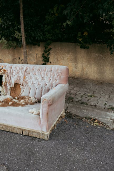A light pink upholstered sofa with a tufted backrest and armrests, positioned outdoors on a paved surface adjacent to a rough concrete wall with visible stains and discoloration. The sofa appears damaged and is covered with a pile of torn fabric and stuffing remnants, indicating it has been discarded or left for rubbish collection. To the right of the sofa, the pavement transitions into uneven stone or concrete blocks that form a small pathway or driveway. Behind the sofa, dense green foliage and leafy branches extend partially over the wall, providing a natural backdrop. The environment suggests an outdoor area possibly near a garden or alleyway where waste materials, including old furniture, are temporarily stored or awaiting private rubbish removal services, such as those offered by Rubbish Collection Marylebone. The lighting is natural, indicating daytime, with no artificial shadows or reflections present.