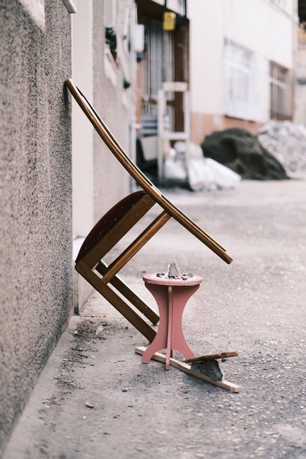 The image shows a narrow outdoor alleyway or passageway filled with various discarded furniture and household items, including a wooden cabinet with ornate detailing, a fabric-upholstered chair, a black office chair, and several pieces of wood and cardboard scattered on the uneven brick ground. The surrounding walls are beige and weathered, with some peeling paint and debris accumulated along their bases. Overhead, a makeshift, partially collapsed canopy of rusty metal pipes and tattered fabric or plastic sheeting provides minimal coverage, casting shadows across the cluttered scene. The environment appears to be an unmanaged waste accumulation area, possibly used for private rubbish collection or on-site clearance, with visible signs of neglect. This scene aligns with the type of bulk waste or furniture removal service that Rubbish Collection Marylebone offers, demonstrating a typical setting where independent rubbish removal services are essential for clearing and disposal of unwanted household items in Marylebone.