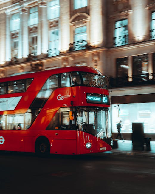 A bright red double-decker bus navigating along a city street at night, with illuminated windows and a digital display indicating the route to Marylebone, part of the Go London transportation network. The bus is in motion, captured with a slight blur that emphasizes its movement through the urban environment. In the background, a historic building with ornate architectural details, large windows, and classical columns is visible, lit softly by ambient street lighting. Pedestrians can be seen walking along the sidewalk beside the bus, some near storefronts with glass display windows. The overall scene suggests a busy, well-lit city center, relevant to urban public transportation and private waste disposal options that may be part of integrated city management or extraregional disposal services like those offered by Rubbish Collection Marylebone. The image maintains a professional, factual tone, focused on urban transit and surroundings, without promotional language.