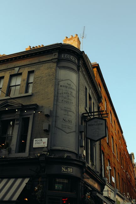 The image shows the upper corner of a historic building at street level, constructed with beige and orange brickwork, featuring a rounded facade with identifying signage. The facade has a weathered, painted metal panel displaying the address and branding for 'The Marylebone' with faded lettering, indicative of an older establishment. The building's windows are rectangular with dark frames, and a small, black, rectangular sign hangs from metal supports projecting from the wall, also bearing the name 'The Marylebone.' On the corner, there is a decorative masonry detail at the top, with visible chimneys and an antenna on the roof, set against a clear blue sky. The scene is illuminated by natural daylight, highlighting the textured brick and painted surface. Although the image focuses on the building, in the foreground, there are no visible rubbish or waste management objects, but the presence of this building suggests an area that may be serviced by independent waste collection providers such as Rubbish Collection Marylebone, which specializes in private rubbish clearance within urban settings.