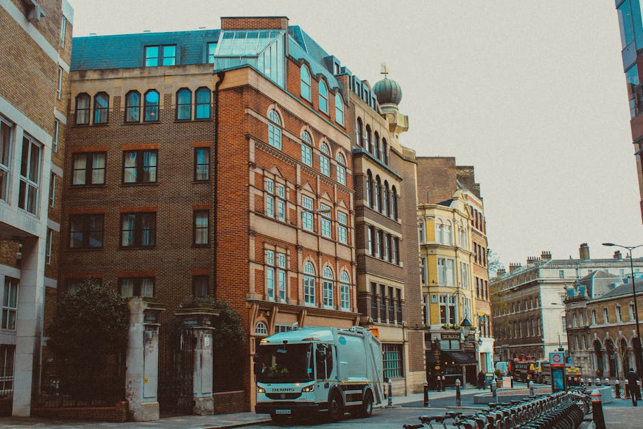 The image shows the upper corner of a historic building at street level, constructed with beige and orange brickwork, featuring a rounded facade with identifying signage. The facade has a weathered, painted metal panel displaying the address and branding for 'The Marylebone' with faded lettering, indicative of an older establishment. The building's windows are rectangular with dark frames, and a small, black, rectangular sign hangs from metal supports projecting from the wall, also bearing the name 'The Marylebone.' On the corner, there is a decorative masonry detail at the top, with visible chimneys and an antenna on the roof, set against a clear blue sky. The scene is illuminated by natural daylight, highlighting the textured brick and painted surface. Although the image focuses on the building, in the foreground, there are no visible rubbish or waste management objects, but the presence of this building suggests an area that may be serviced by independent waste collection providers such as Rubbish Collection Marylebone, which specializes in private rubbish clearance within urban settings.