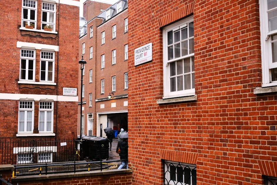 A narrow urban alleyway flanked by red brick residential buildings with white-framed sash windows, some with flower pots on the sills. In the foreground, part of a brick wall and metal railing are visible, leading to a driveway where two individuals, dressed in dark outdoor clothing, are seen handling black wheelie bins. The scene includes a street sign indicating Portland Square and Providence Court W1, and a vintage-style street lamp affixed to one building. The background shows additional multi-storey brick buildings with small windows, creating a typical London residential setting. The alley is clean, with some visible yellow barriers on the pavement near the bins, suggesting routine rubbish collection or private waste disposal. The overall atmosphere is daytime, with natural light illuminating the brick textures and urban environment, relevant to independent rubbish collection services in Marylebone.
