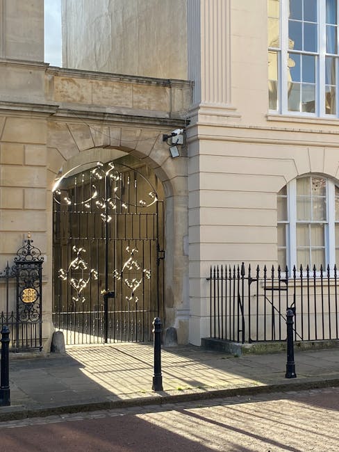 Photograph of the exterior of a historic building with a grand arched entrance framed by a stone facade and tall, white, rectangular windows. The dark metal gate within the arch features decorative, ornate patterns and is slightly open, casting striped shadows on the pavement due to sunlight. To the left of the gate, there is a black wrought iron fence with intricate scrollwork, and a small decorative plaque or emblem mounted on the fence. The building's architecture includes classical elements such as pilasters and detailed cornices. The scene is lit by natural daylight, creating a high-contrast environment with shadows cast across the pavement and entrance area, suggesting a quiet street setting. This image, associated with professional rubbish collection services, visually reflects the importance of maintaining the cleanliness and aesthetic appeal of historic property exteriors through proper waste handling and clearance.