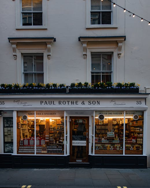 The image depicts the storefront of Paul Rothe & Son, a delicatessen and grocery shop located at number 35. The shop's large display windows on either side of a central wooden door showcase an array of goods, including jars, bottles, and packaged foods, arranged neatly on shelves. The exterior is painted white with black trim at the bottom, and the shop's name is displayed on a sign above the windows in elegant, traditional lettering. Above the store, there are two residential-style windows with white frames and decorative trim, one of which has a windowsill decorated with yellow and purple flowers in a flower box. String lights are hung across the upper part of the scene, adding a warm atmosphere. The overall scene is set on a paved sidewalk in daylight, suggesting a quiet street environment, which could relate to the context of commercial premises that may require efficient waste disposal or regular rubbish collection. The image offers a clear view of a traditional shopfront, potentially benefitting from services such as independent rubbish removal for small businesses.