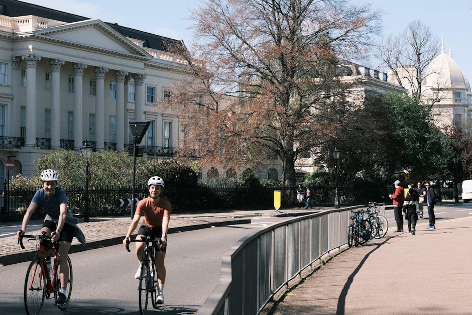The image depicts a city street scene with two cyclists wearing helmets riding on the roadway in the foreground, one in a navy blue shirt and the other in a brown T-shirt, both appearing to enjoy a leisurely ride. Behind them, there is a row of parked bicycles secured to a metal bike rack on a paved sidewalk. Several pedestrians are visible further back, standing and walking near the curb, some engaged in conversation while others are heading along the sidewalk. The background features an elegant, historic white building with tall columns, decorative moldings, and large windows, set behind a black wrought-iron fence and lush greenery, including shrubbery and trees, one of which has a bare, textured trunk and branches suggesting it's late autumn or winter. To the right, a distinctive building with a dome-shaped roof is partially visible behind the trees. The sky above is mostly clear, with natural daylight illuminating the scene, creating a calm urban atmosphere. The image aligns with professional waste management and rubbish removal themes by illustrating an environment where proper waste handling supports a clean and organized cityscape, as represented by the overall neat streets and surroundings, subtly referencing the importance of municipal and private rubbish collection services. Rubbish Collection Marylebone provides essential services to maintain such environments, facilitating alternative waste handling and on-site clearance solutions in urban areas.
