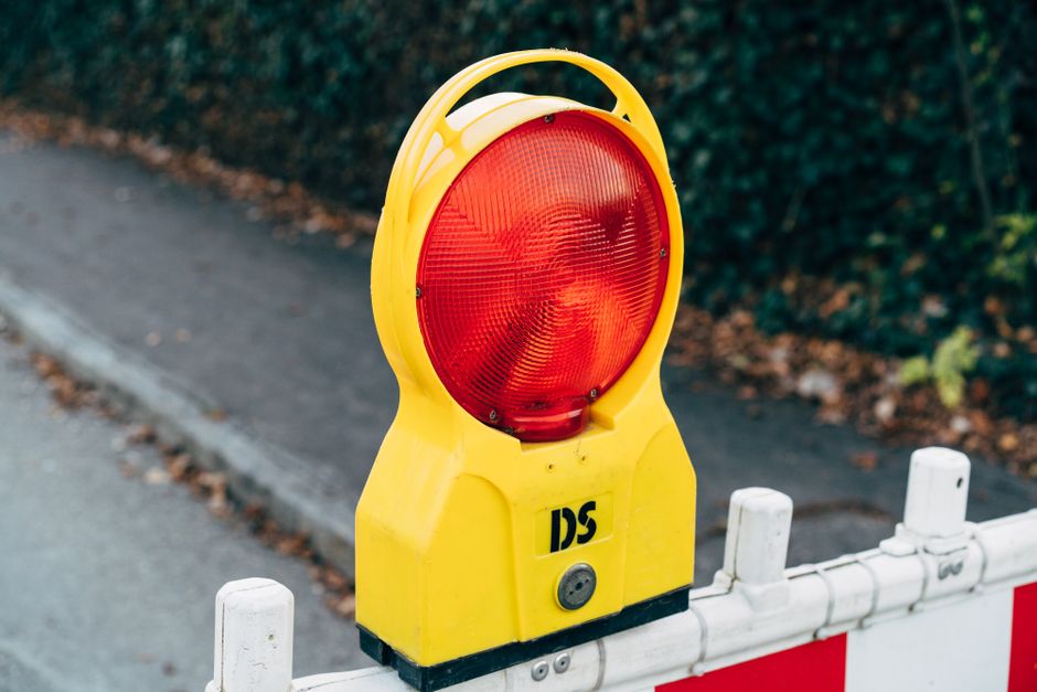 A close-up view of a portable yellow warning light with a large, circular, red reflective lens mounted on a black base. The yellow casing has a textured finish and a sturdy handle integrated into the top. The warning light is positioned in front of a temporary road barrier with a red and white reflective pattern, indicating a construction or work zone area. The background shows a section of pavement with scattered brown leaves along the curb, and dense green foliage beyond, suggesting an outdoor setting. The lighting appears natural, with no shadows indicating diffuse daylight. This setup is typical for site safety and traffic management, often associated with construction or roadwork activities where clear marking for alternative waste handling or site clearance is necessary, as managed by companies like Rubbish Collection Marylebone.