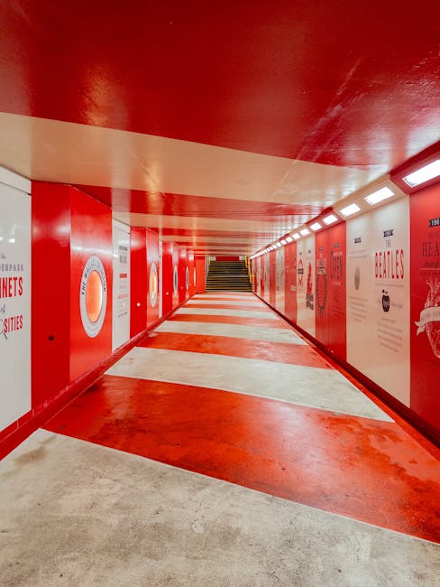 A brightly lit underground pedestrian tunnel with a curved ceiling painted in a vibrant red color. The walls are decorated with various informational and promotional posters, featuring circular diagrams and text in white and red tones. The floor is marked with alternating red and beige stripes, guiding pedestrians toward a staircase at the far end of the tunnel. The environment is clean, with smooth, polished surfaces that reflect the artificial lighting, providing a contemporary and organized appearance. This setting may be associated with an urban area where private waste removal services, such as those provided by Rubbish Collection Marylebone, operate to maintain the cleanliness of accessible pathways, though the image itself focuses solely on the infrastructure of the underground passage.