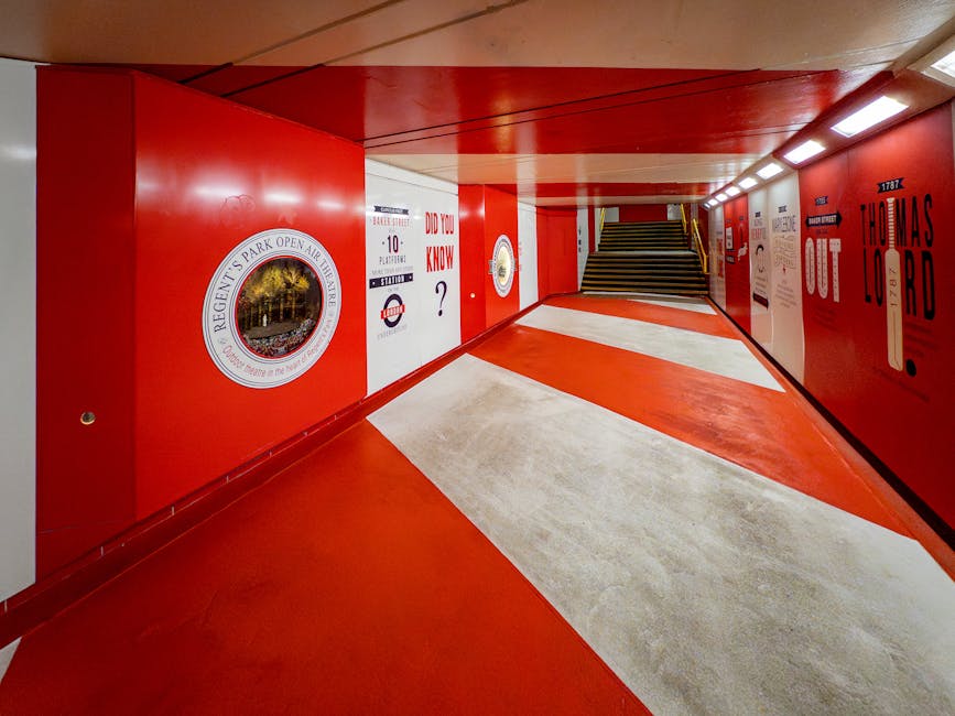 A brightly lit underground pedestrian tunnel with a curved ceiling painted in a vibrant red color. The walls are decorated with various informational and promotional posters, featuring circular diagrams and text in white and red tones. The floor is marked with alternating red and beige stripes, guiding pedestrians toward a staircase at the far end of the tunnel. The environment is clean, with smooth, polished surfaces that reflect the artificial lighting, providing a contemporary and organized appearance. This setting may be associated with an urban area where private waste removal services, such as those provided by Rubbish Collection Marylebone, operate to maintain the cleanliness of accessible pathways, though the image itself focuses solely on the infrastructure of the underground passage.