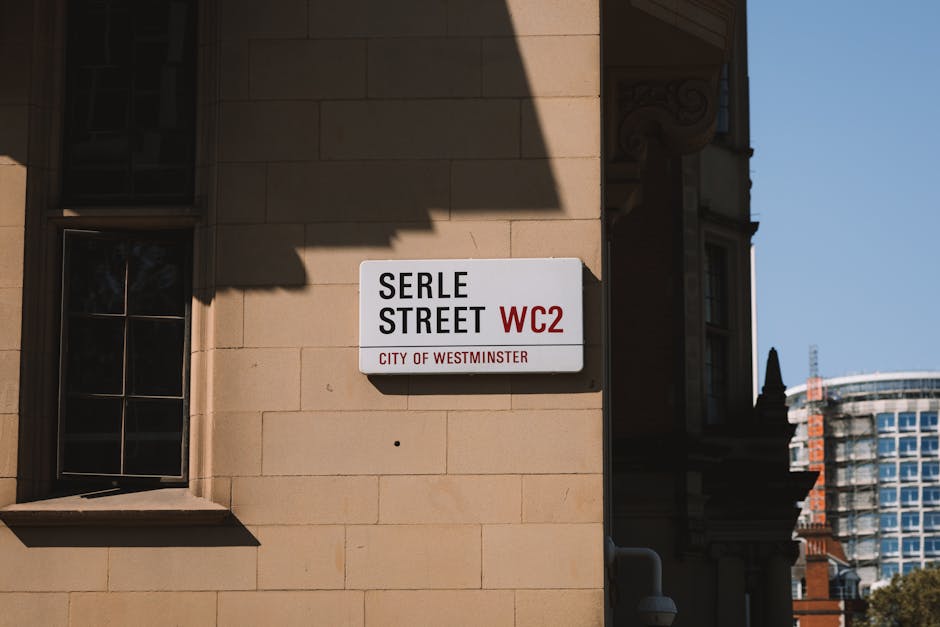 A rectangular street sign mounted on a beige stone building wall indicating 'Serle Street' with the postcode WC2 and the label 'City of Westminster' in small red text beneath. The sign has a white background with black and red text, positioned slightly to the right of a window with a black frame and multiple panes, which casts a shadow on the wall. The scene is captured during daylight, with a clear blue sky visible in the background. Adjacent to the building, the background includes a modern high-rise building with glass windows and a rounded structure, suggesting an urban setting. The photograph's lighting emphasizes the texture of the stone wall and the sign, situating it within a city environment that may be associated with areas serviced by independent waste removal companies like Rubbish Collection Marylebone, which provides alternative waste handling options outside standard municipal rubbish collection.