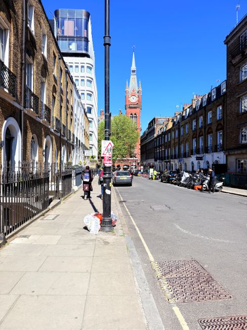 A rectangular street sign mounted on a beige stone building wall indicating 'Serle Street' with the postcode WC2 and the label 'City of Westminster' in small red text beneath. The sign has a white background with black and red text, positioned slightly to the right of a window with a black frame and multiple panes, which casts a shadow on the wall. The scene is captured during daylight, with a clear blue sky visible in the background. Adjacent to the building, the background includes a modern high-rise building with glass windows and a rounded structure, suggesting an urban setting. The photograph's lighting emphasizes the texture of the stone wall and the sign, situating it within a city environment that may be associated with areas serviced by independent waste removal companies like Rubbish Collection Marylebone, which provides alternative waste handling options outside standard municipal rubbish collection.